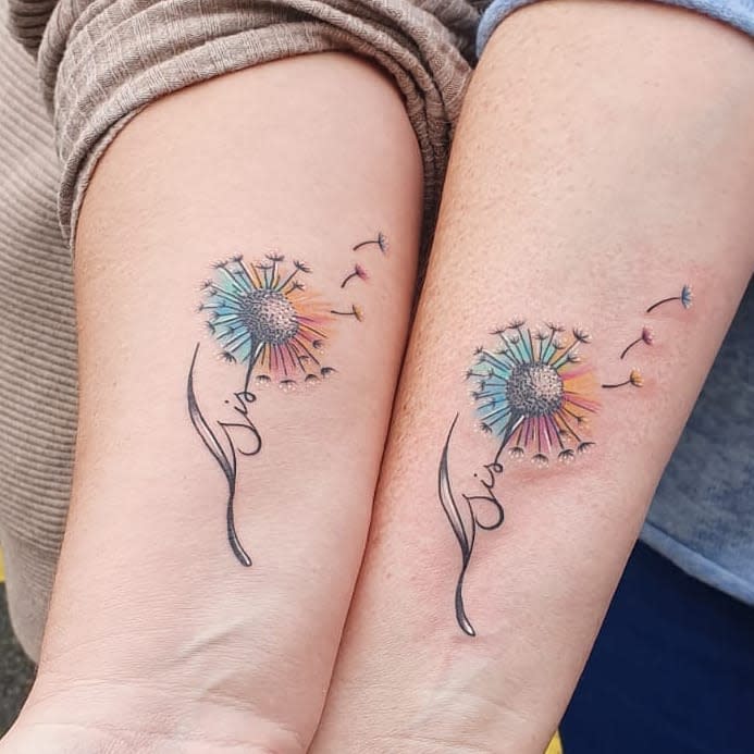 Two arms with matching colorful dandelion tattoos, seeds blowing away, and cursive text